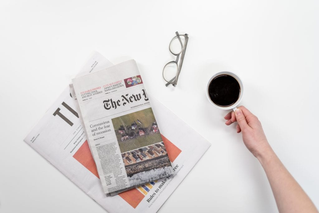 A hand reaches for a cup of coffee beside two newspapers and a pair of glasses