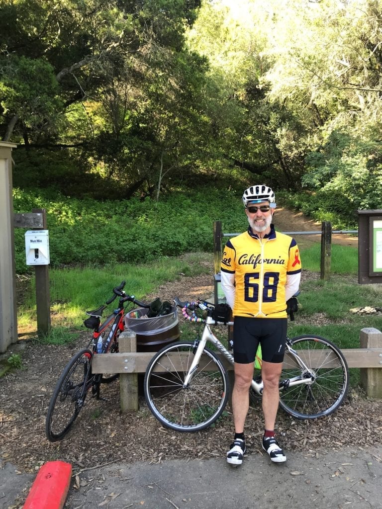 Harry Stark stands with his bike in a Cal jersey with trees and a hiking path behind him. 