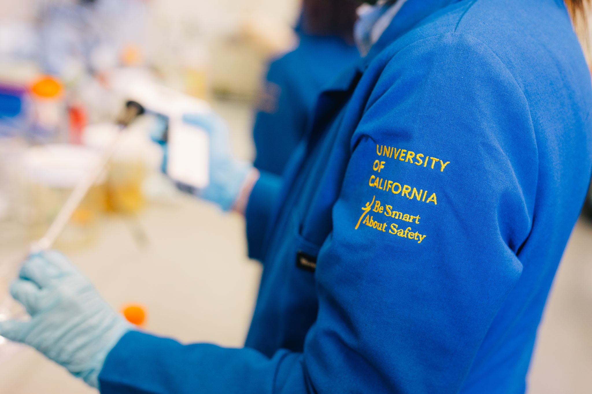 A researcher in a blue University of California lab coat holds a pipette