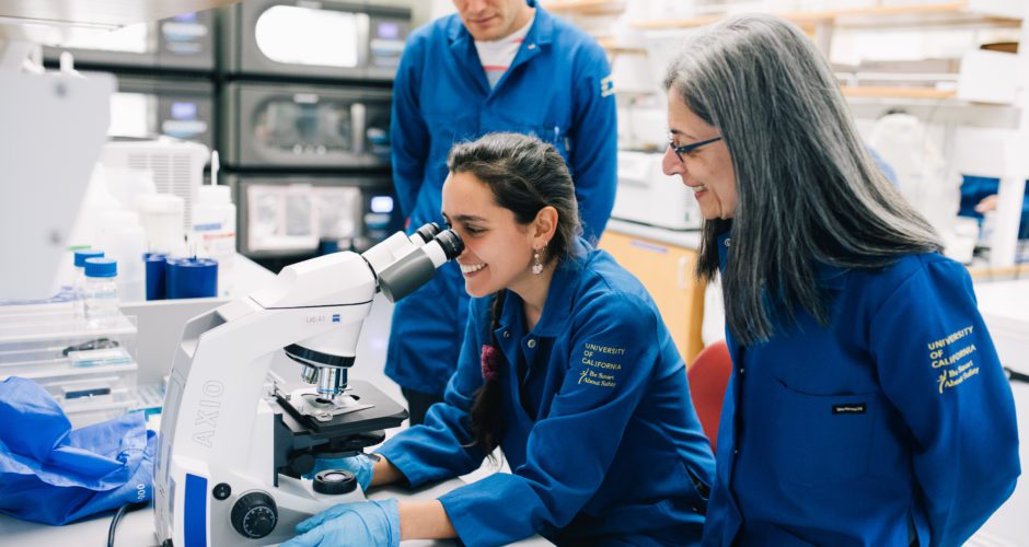 Two researchers stand in blue University of California lab coats and watch another researcher looking into a microscope.