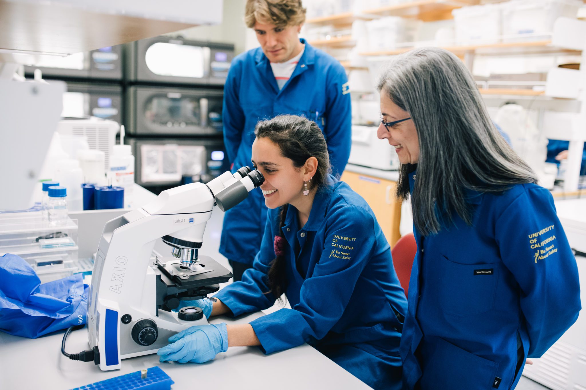 Two researchers stand in blue University of California lab coats and watch another researcher looking into a microscope.