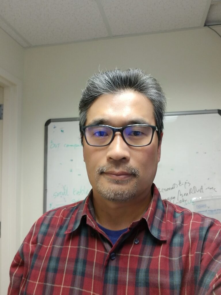 Headshot of Justin Choi in glasses and plaid shirt standing in front of a white board.