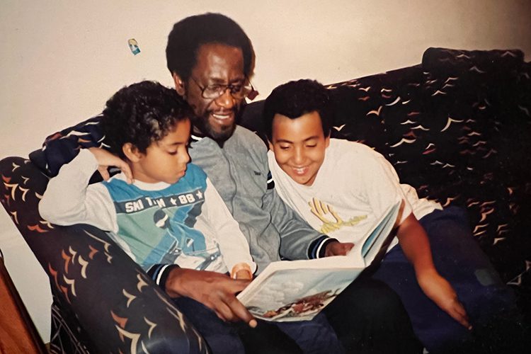 A slightly older photograph of a Black man wearing glasses sitting on a couch reading a book to two Black boys. 