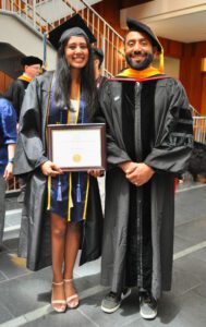 Two people standing in an academic building courtyard in full regalia. One is holding a framed diploma. 