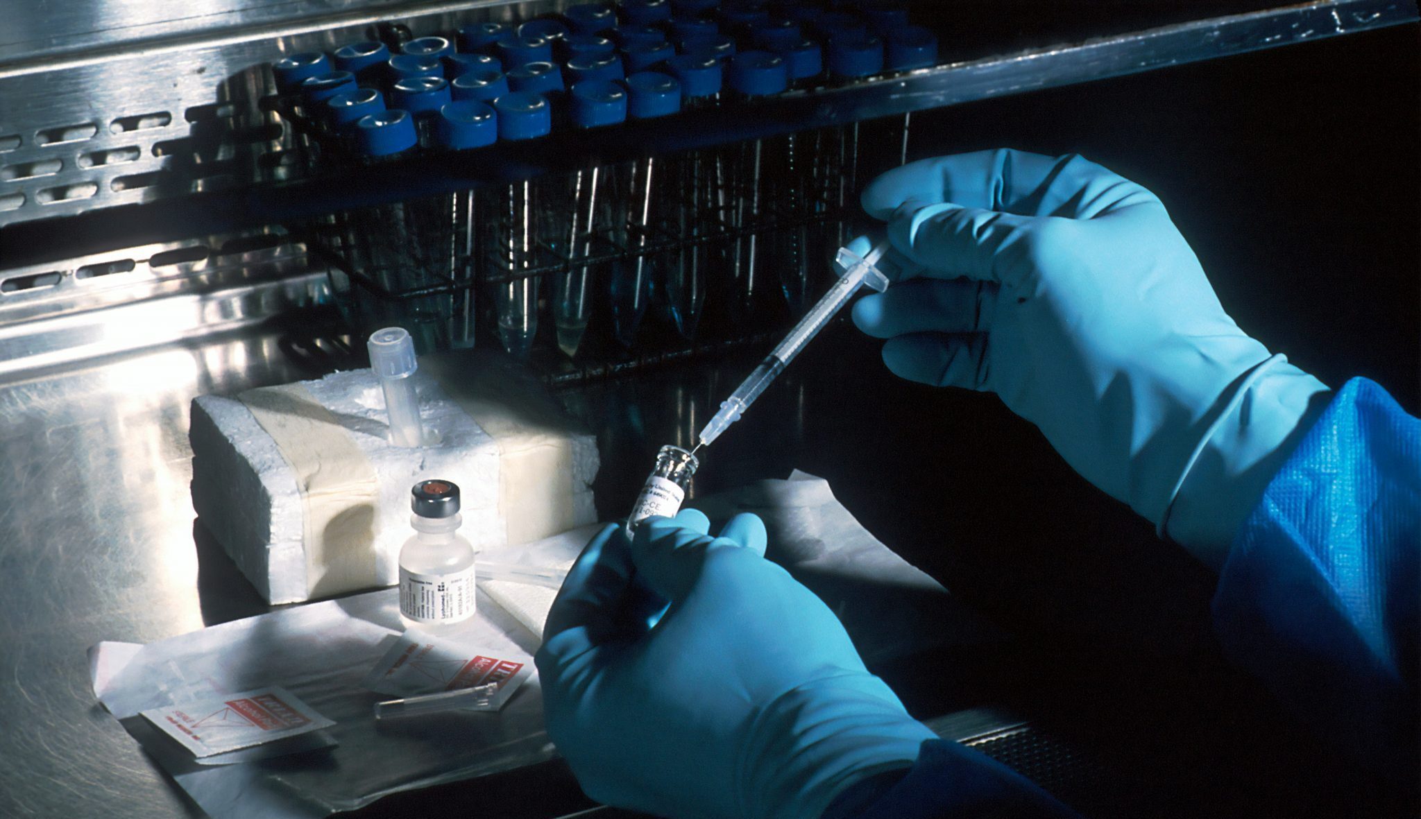 A pair of hands in blue sterile gloves inserting a syringe into a vial.