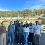 A group of people from the Lucas lab stand outside in front of a blue sky and green hills.